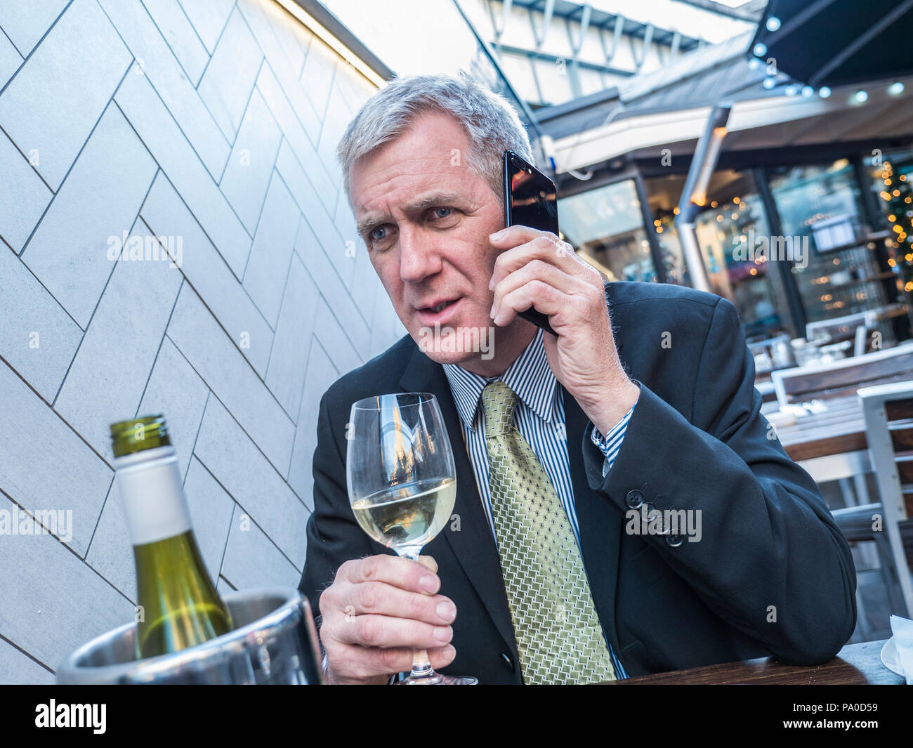 Imprenditore con bicchiere di vino audizione cattive notizie cerca preoccupato di parlare in ascolto sul suo iPhone telefono cellulare al ristorante alfresco bar Foto Stock