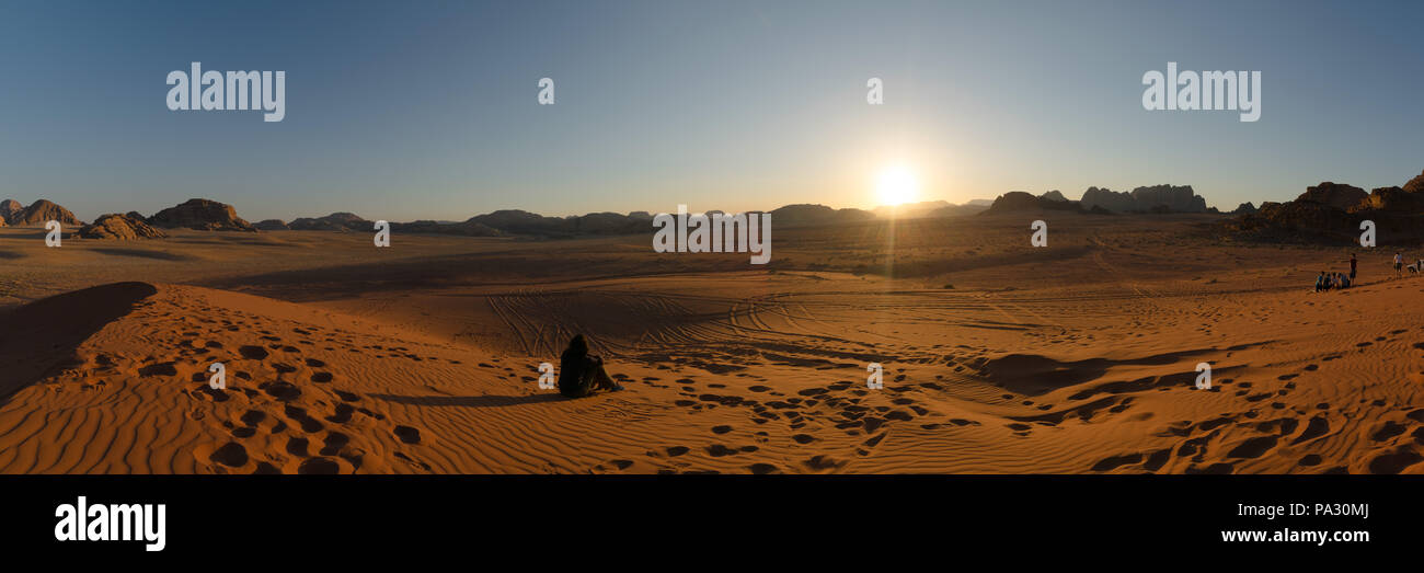 Una turista femminile siede su una duna di sabbia di fronte al sole tramontare su formazioni rocciose nel popolare luogo di trekking deserto Wadi Rum, Giordania Foto Stock