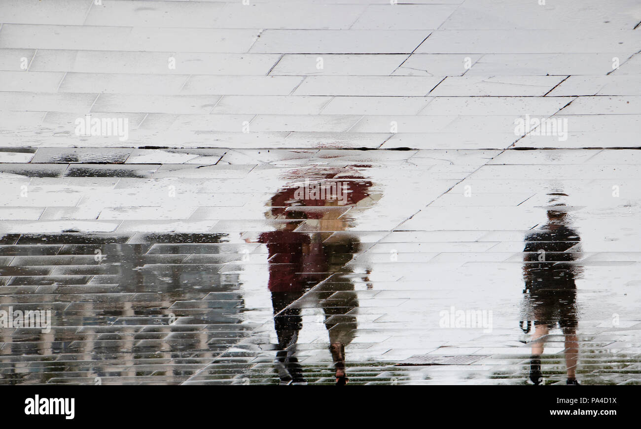 Sfocata riflessione silhouette di una giovane coppia camminare sotto ombrellone rosso e un uomo a camminare da solo e a basso angolo di visione, nella pozza , in primavera piovosa Foto Stock