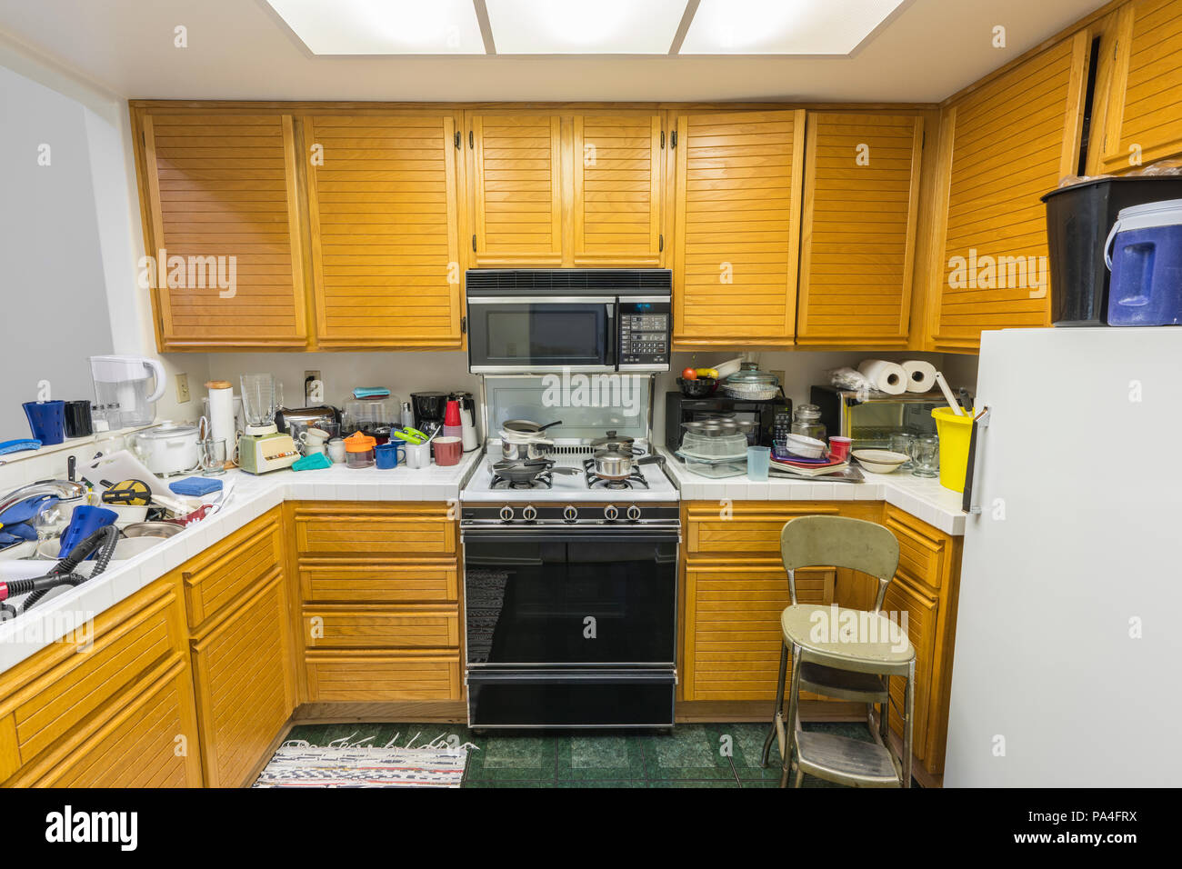 Confuso il vecchio appartamento cucina con armadi in legno di quercia, tile countertops, fornello a gas a pavimento verde e pile di piatti. Foto Stock