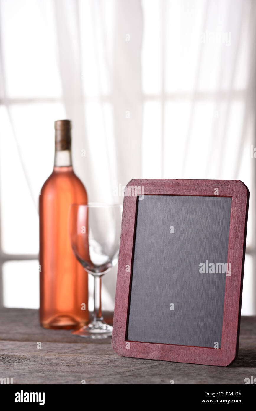 Una bottiglia di vino arrossire con un menu vuoto bordo, su una tavola in legno rustico di fronte a una finestra. Perfetto per un vino o Menu Degustazione Vini annuncio. Foto Stock