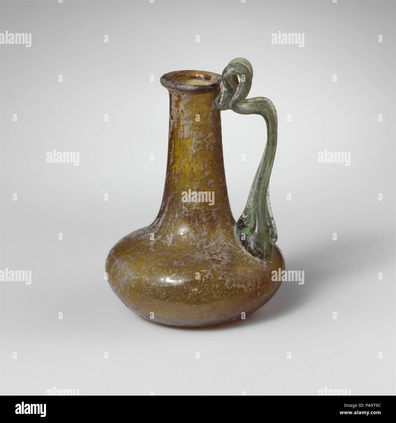 Caraffa di vetro. Cultura: Romano. Dimensioni: complessivo: 3 5/16 in. (8.4Cm) Diam.: 2 1/4 x 13/16 in. (5,7 x 2,1 cm). Data: 1a-2a secolo D.C.. Traslucido giallo bruno, con traslucido luce maniglia verde. Cerchione piegato fuori, sopra e in, appiattita con superficie superiore; collo cilindrico, si espande verso il basso; squat, corpo a bulbo; fondo concavo; ampia maniglia a fascia applicata in tampone di grandi dimensioni con due griffe esterna alla parte superiore del corpo, redatto, poi trasformato in verticalmente e fissato nel collo sotto il cerchione con ansa verticale come appoggio per il pollice al di sopra. Intatti; alcune bolle pinprick; assopimento e debole resistenza agli agenti atmosferici di calcaree con s Foto Stock