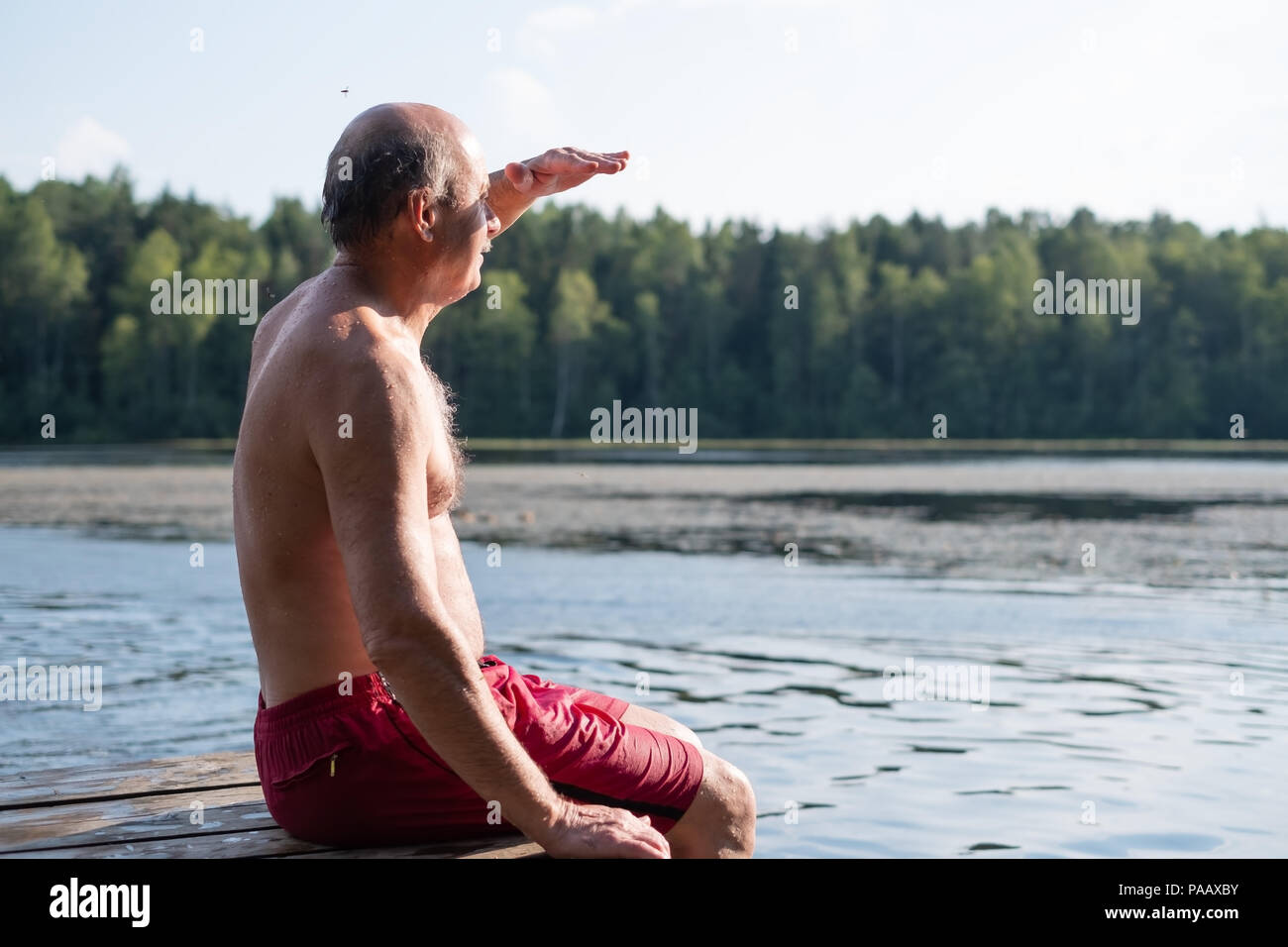 Bel uomo maturo seduta sulle sponde di un lago in una posizione soleggiata e tranquilla giornata, seduto su di un molo di legno appoggiata in villaggio Foto Stock