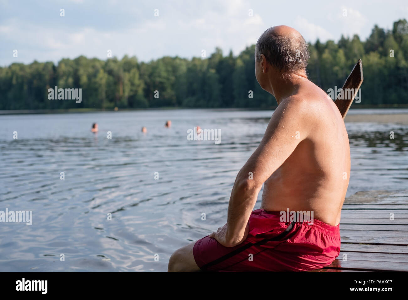 Bel uomo maturo seduta sulle sponde di un lago in una posizione soleggiata e tranquilla giornata, seduto su di un molo di legno appoggiata in villaggio Foto Stock