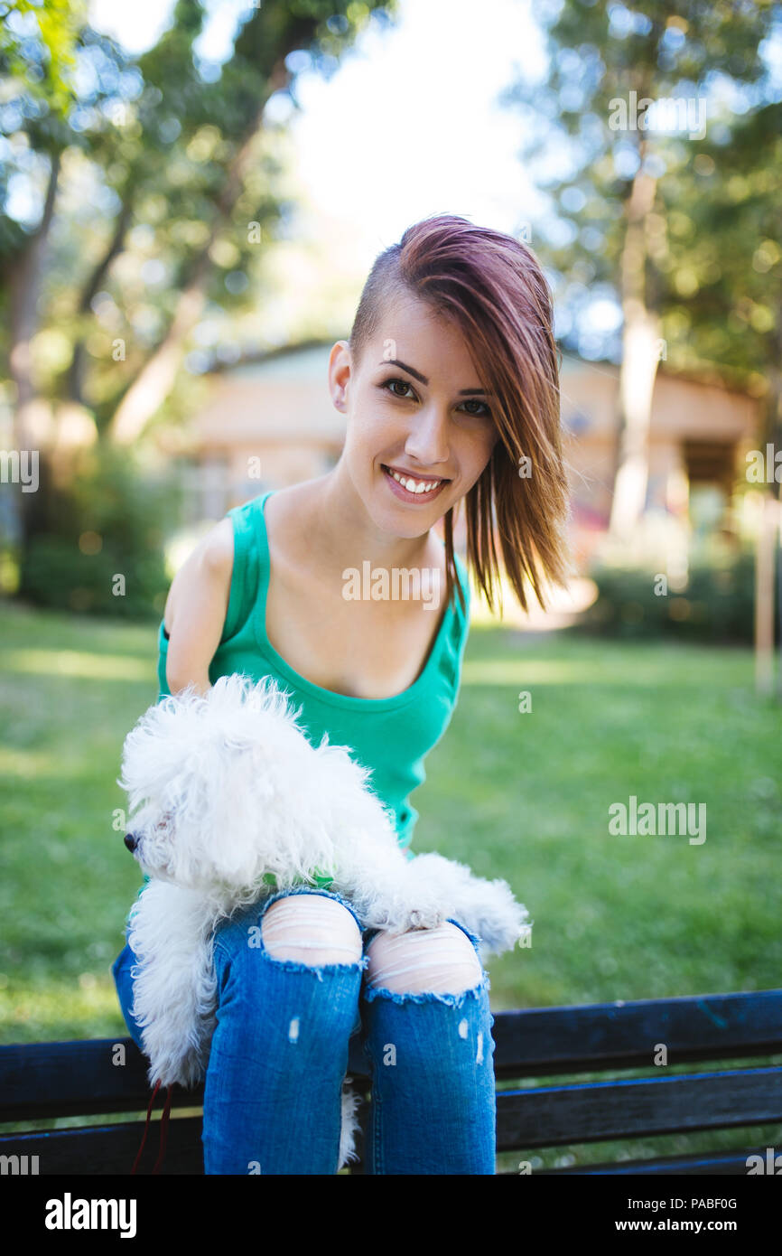 Ritratto di bella non valido sorridente ragazza nata senza arti superiori mantenendo il suo bianco piccolo cucciolo e sedersi sulla panchina nel parco. Foto Stock