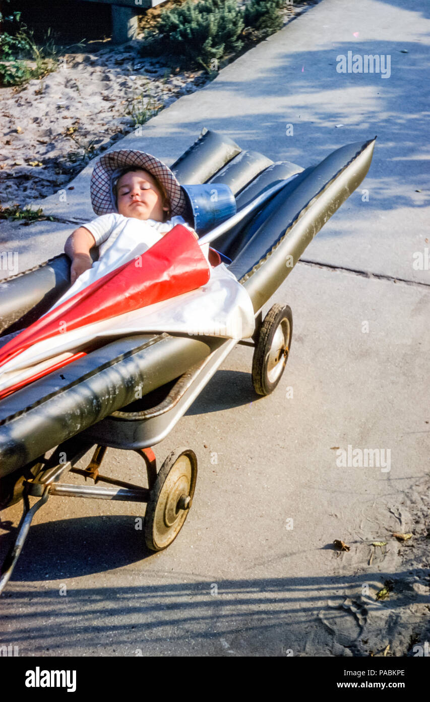 Ragazzo piccolo che indossa cappello veloce addormentato sdraiato su un  lilo gonfiabile in un carro da spiaggia con ombrellone e secchio o secchio,  Breezy Point, Long Island, Queens, New York City, USA