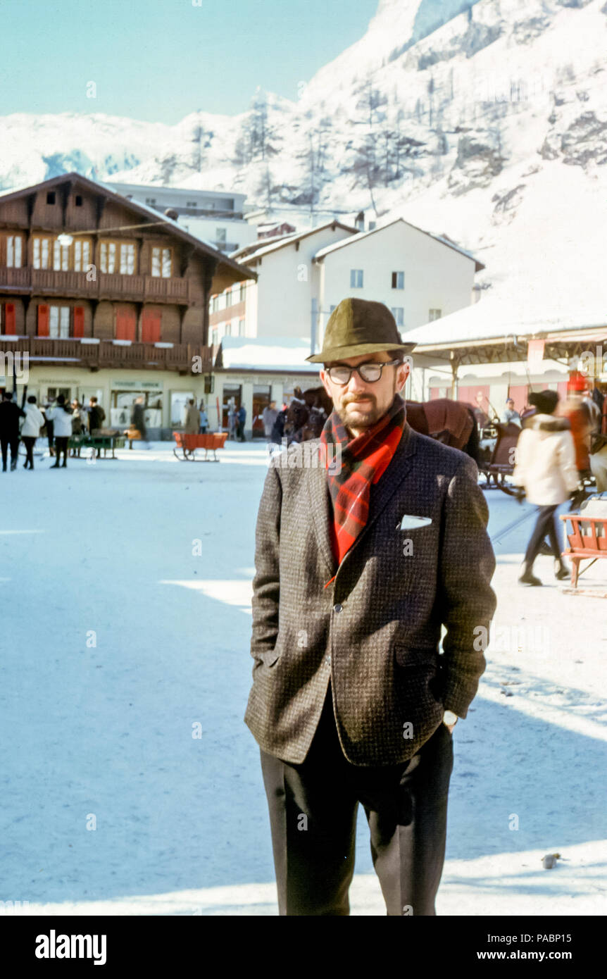 Uomo con barba e occhiali con giacca in tweed e cappello Trilby in piedi nella piazza della stazione sciistica in inverno, Zermatt, Svizzera negli anni '60 Foto Stock