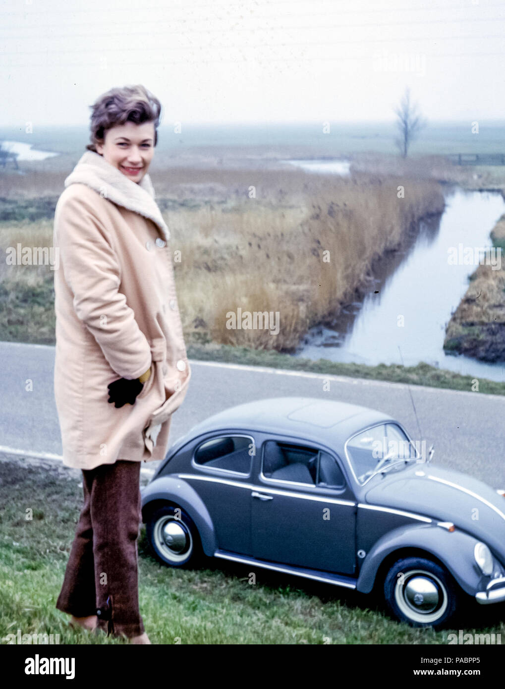 Bella donna che indossa un cappotto invernale in piedi su di una diga olandese con Beetle auto parcheggiata a bordo strada, Olanda, Paesi Bassi, Europa negli anni '60 Foto Stock