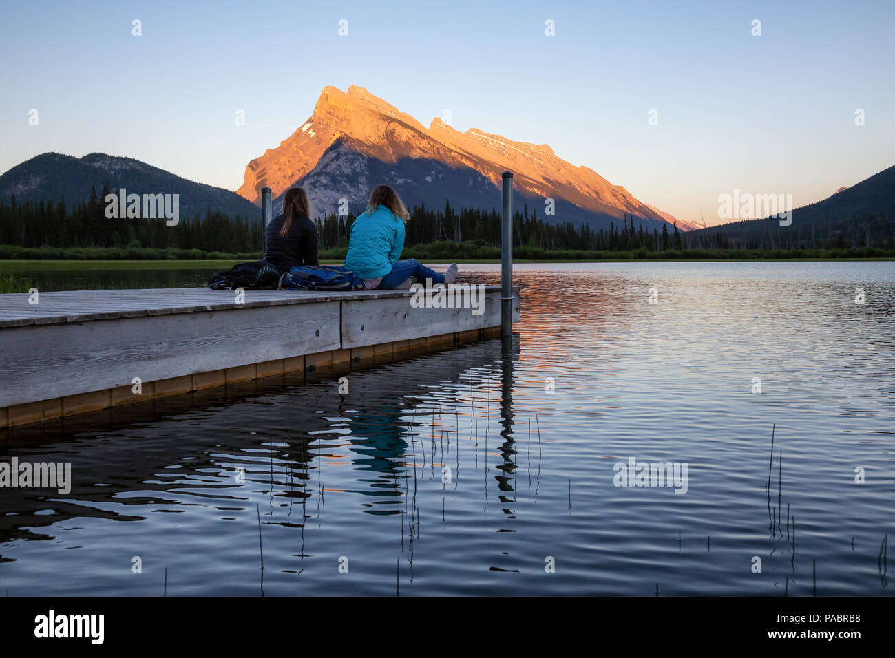 Giovane amici stanno godendo della splendida estate tramonto su un dock in legno dall'acqua. Preso in laghi Vermiglio, Banff, Alberta, Canada. Foto Stock