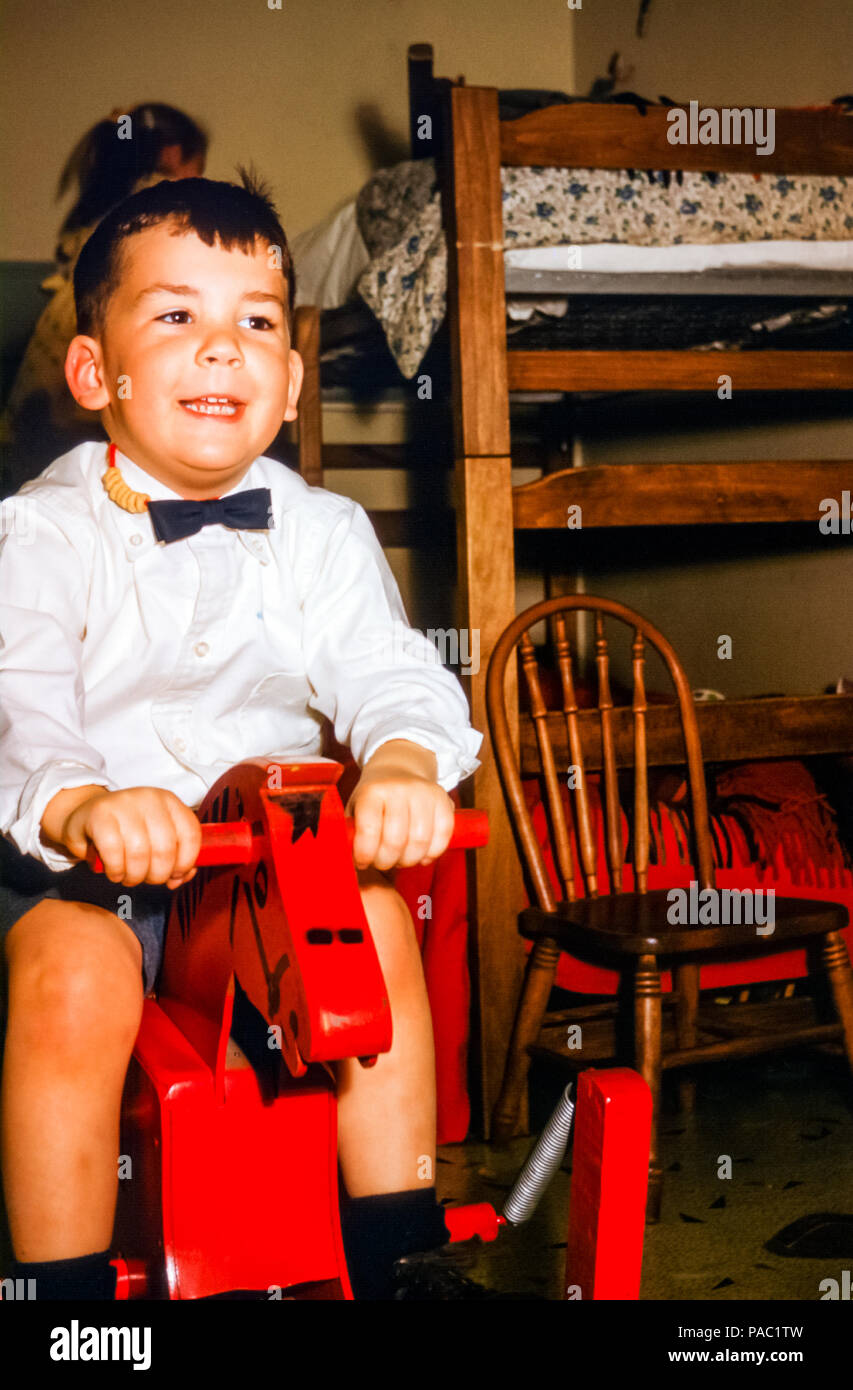 Ragazzo sorridente che indossa una camicia bianca e una cravatta ad arco cavalcare un giocattolo di legno a cavallo in una camera da letto per bambini Con letti a castello negli Stati Uniti negli anni '60 Foto Stock