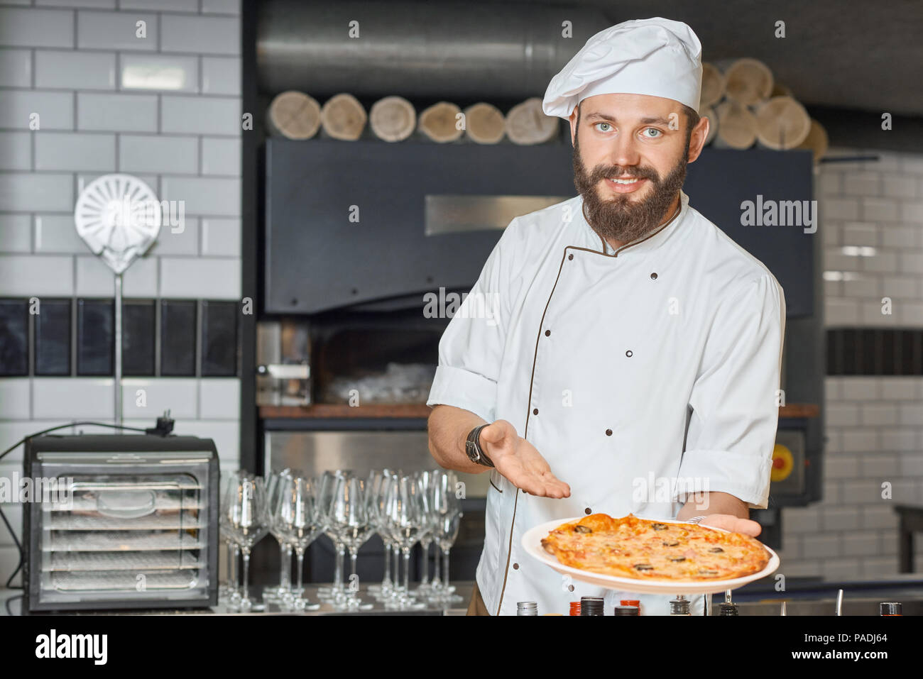 Felice pizzaiolo dimostrando sfornato fresco pizza deliziosa posa sulla  grande lastra. Indossando white chef's tunica lavorando su ristorante  cucina con forno, legnami in legno, bicchieri di vino in piedi sul  background Foto