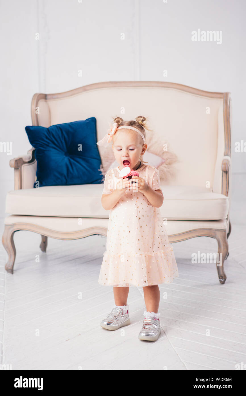 Compleanno di bambini. divertente due-anno-vecchia ragazza caucasica in abito rosa permanente per la camera da letto di casa sullo sfondo del lettino e del mangiare la torta holding des Foto Stock