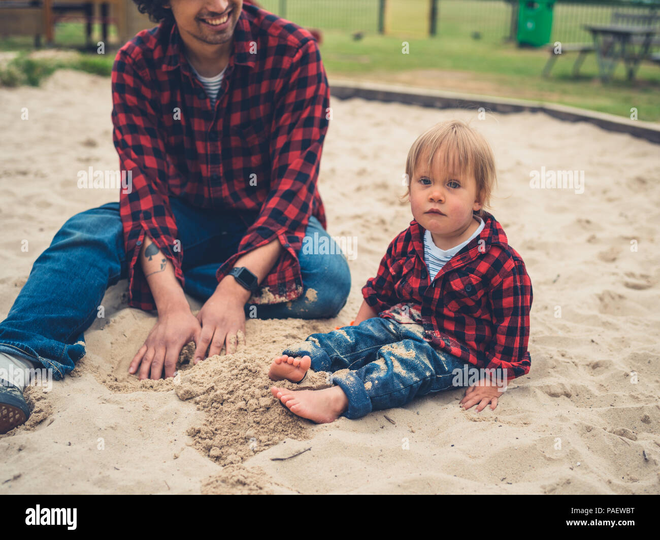 Un giovane padre e suo figlio toddler indossando abiti identici sono seduti in una buca di sabbia al parco giochi Foto Stock