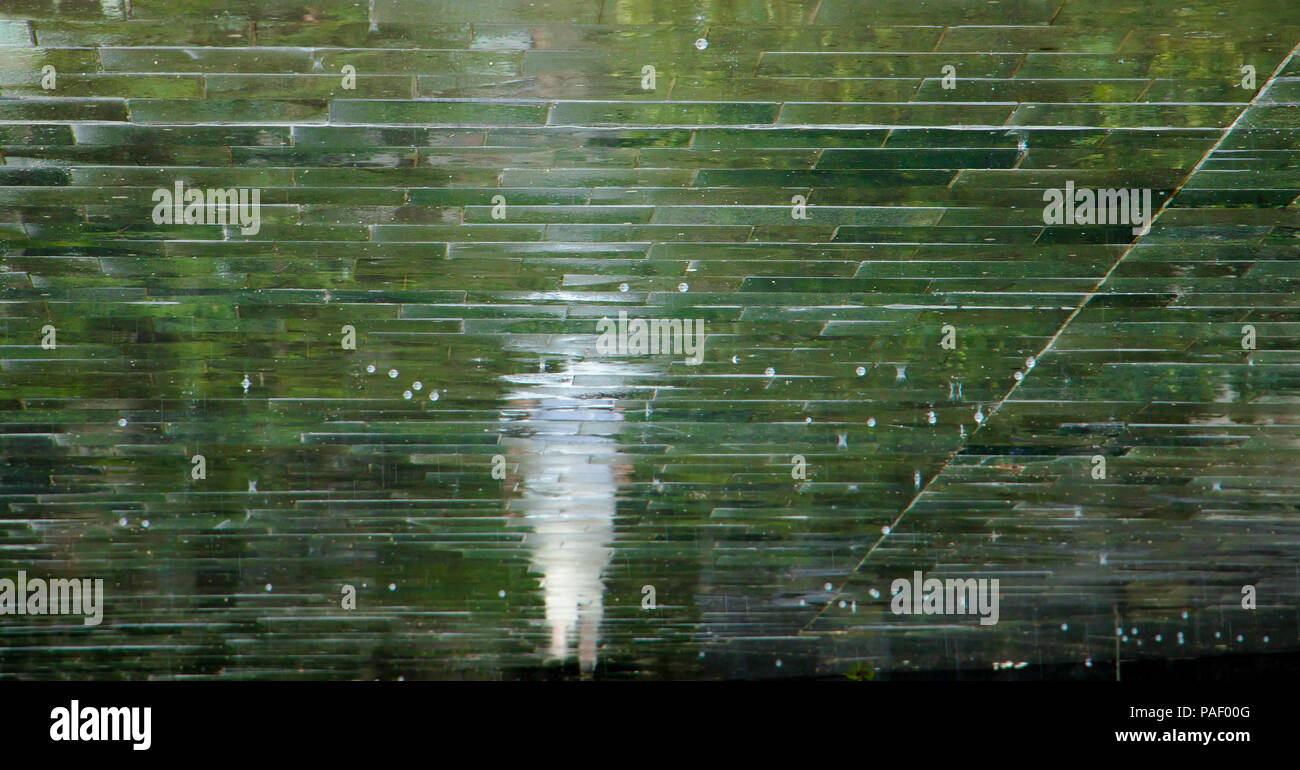 Sfocata riflessione silhouette di una persona che cammina in una piovosa percorso modellato in una città parco verde in estate calda Foto Stock