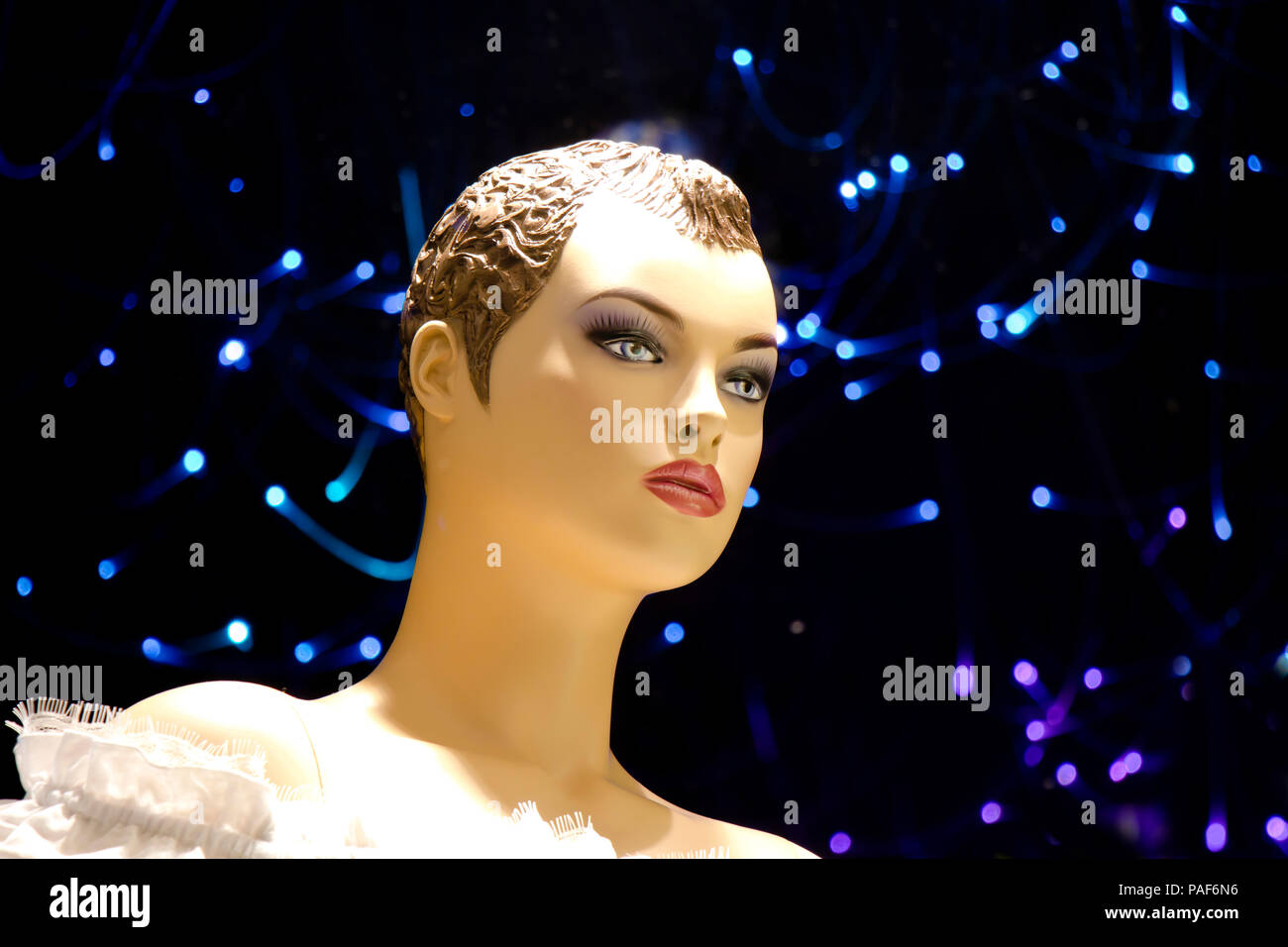 In prossimità di una femmina manichino bambola con breve capelli ricci visualizzato n la vetrina del negozio con sfocate luci blu sullo sfondo , vista di profilo Foto Stock