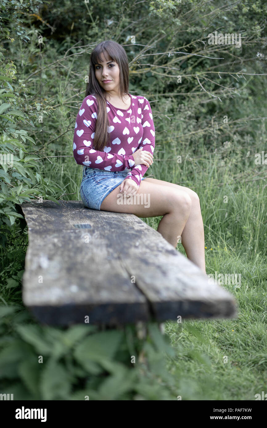 Lo spagnolo donna americana seduta su una panca in legno nel Cambridgeshire countrside. Indossando una susina sommità colorata con il cuore e gli shorts in denim blu. Foto Stock