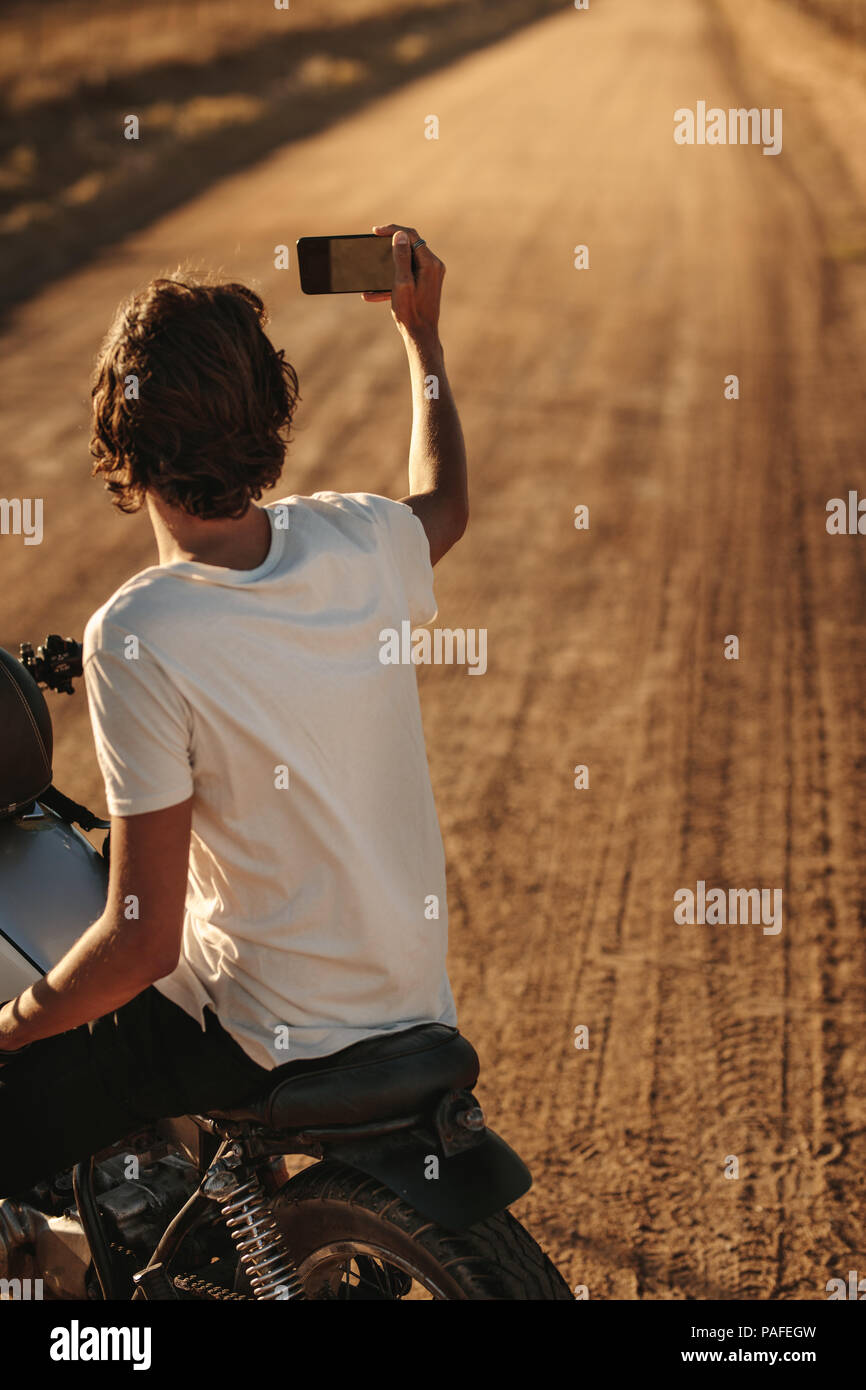 Vista posteriore del giovane uomo prendendo selfie con il suo smart phone mentre è seduto sulla sua moto sulla strada di campagna. Bike Rider catturare i ricordi del suo countr Foto Stock