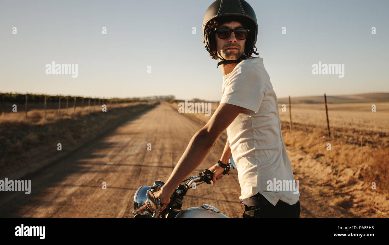 Giovane uomo che indossa un casco e occhiali da sole in piedi con la sua bicicletta e guardando indietro sulla strada rurale. Maschio di bici su strada di campagna. Foto Stock