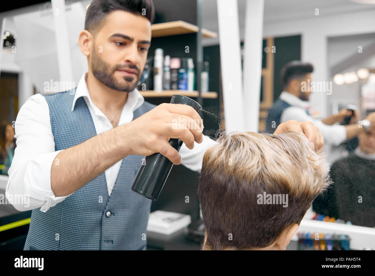 Fare nuova acconciatura per modello maschile mediante fissaggio rigido lacca. Parrucchiere classico che indossa una camicia bianca, gilet grigio. Lavorando in specchio anteriore in beaty salon. Prodotti per la cura dei capelli su sfondo. Foto Stock