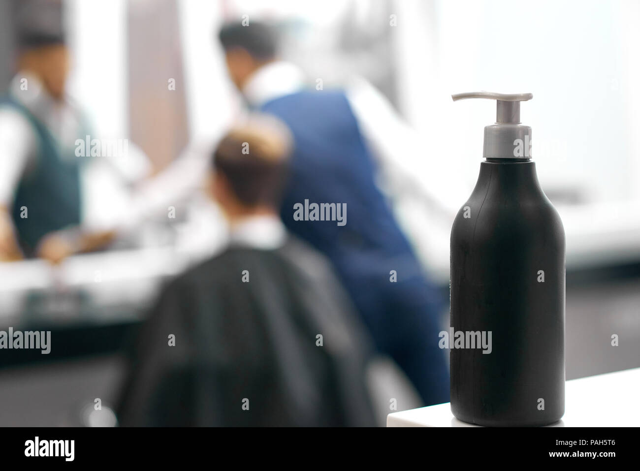 Foto di nero la bottiglia di plastica con erogatore compreso cura dei capelli prodotto. In piedi sul bianco superficie immediatamente in un barbiere, beaty salon. Parrucchiere a lavorare con il cliente nei pressi di specchio sul background. Foto Stock