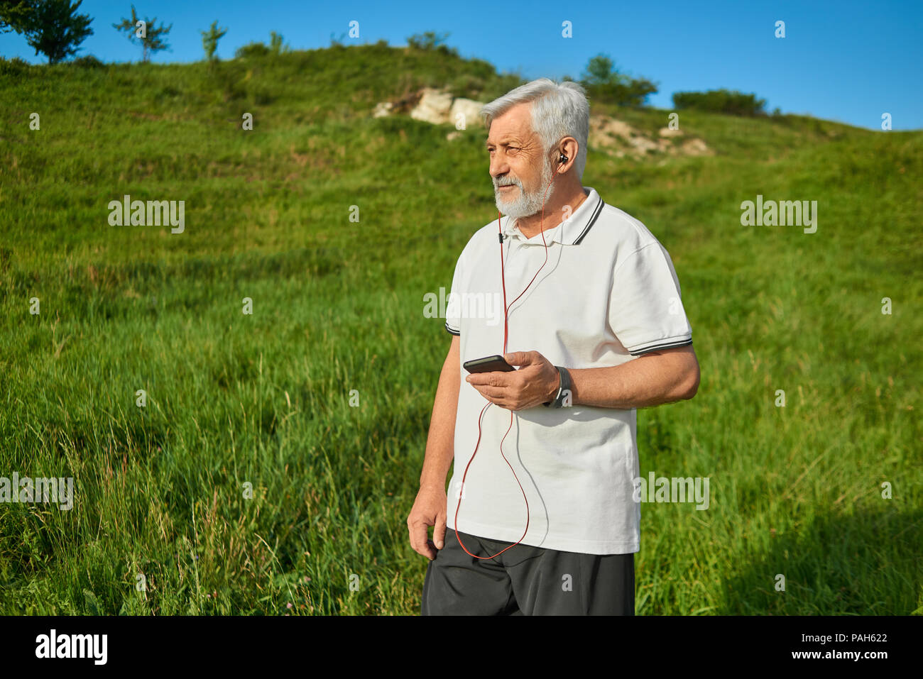Il vecchio uomo mantenendo il telefono cellulare, in piedi sul campo guardando a lato. Ascolto di musica con cuffie rosso. Indossando il bianco classico Polo shirt scuro con strisce blu,pantaloni neri, sneakers. Stile di vita sportiva. Foto Stock