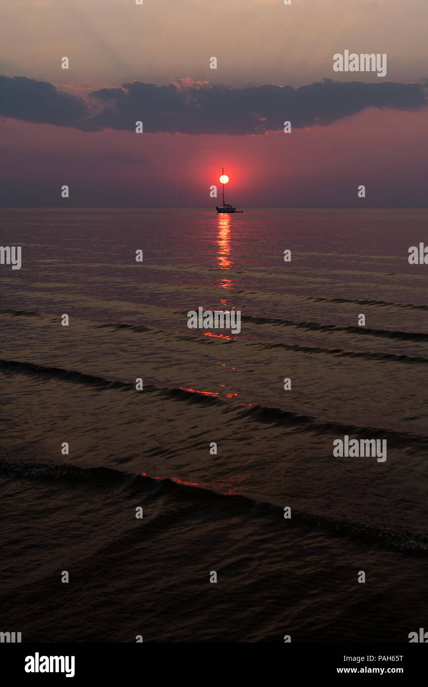 Il sole scende tra le onde del mare contro lo sfondo di yacht e nuvole all'orizzonte Foto Stock