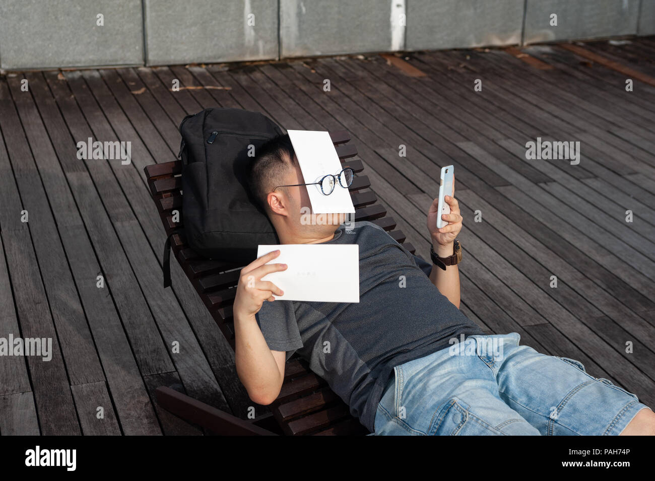 21.07.2018, Singapore, Repubblica di Singapore, in Asia - un uomo che ha il suo volto coperto guarda al suo telefono come egli è disteso su un lettino di Marina Bay. Foto Stock