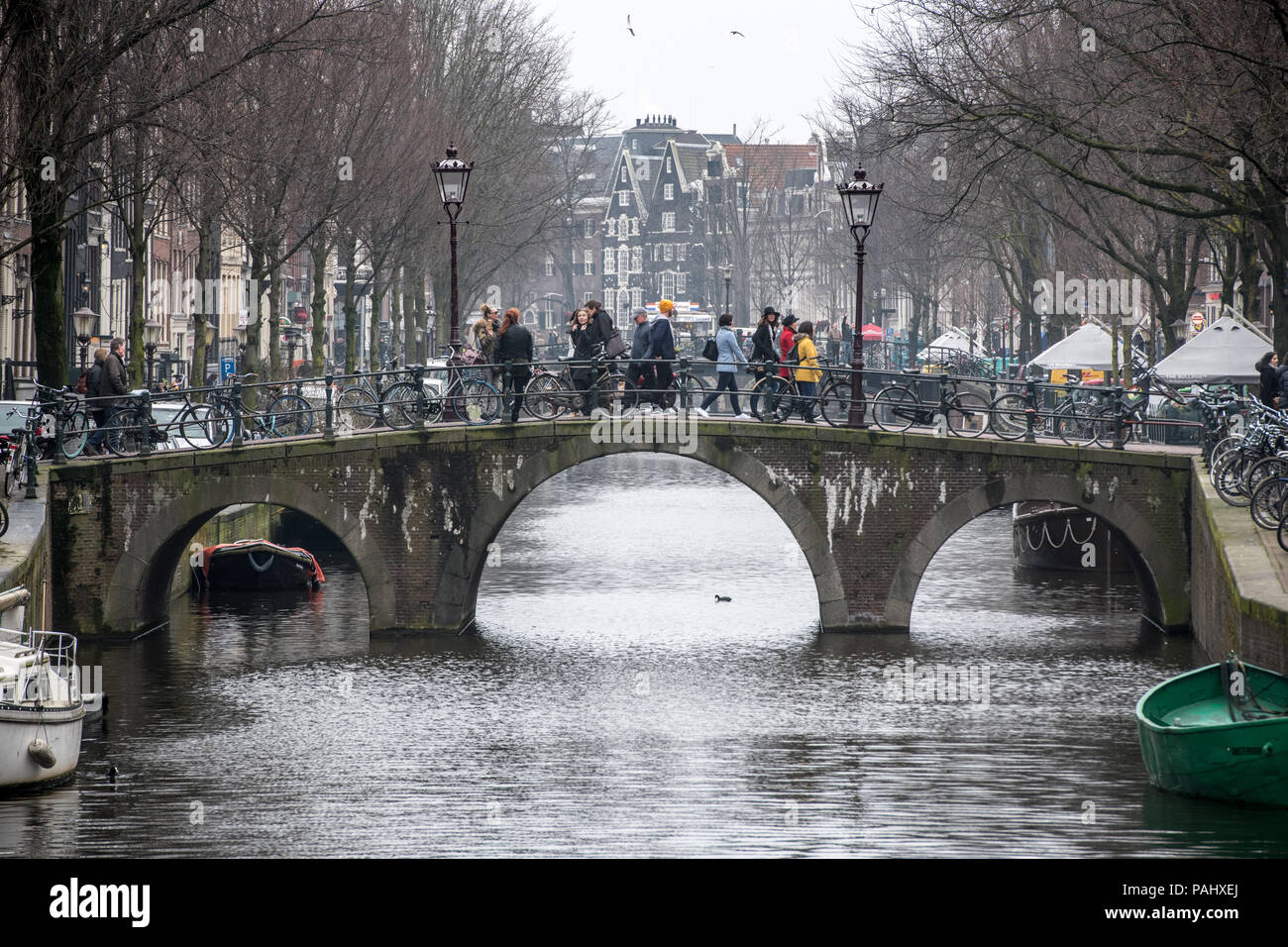 Gruppo di individui utilizzare ponte per attraversare il canale che attraversa la città di Amsterdam con la storica Canal case in background, Paesi Bassi.. Foto Stock