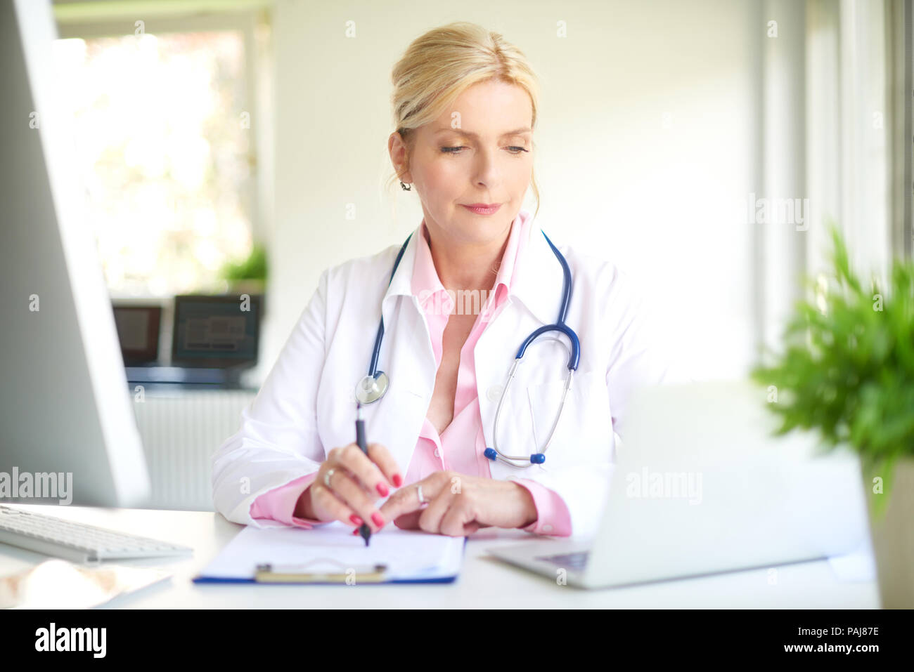 Sorridente anziani medico donna seduta al medico e camera utilizzando il computer portatile e mentre la scrittura di diagnosi. Foto Stock