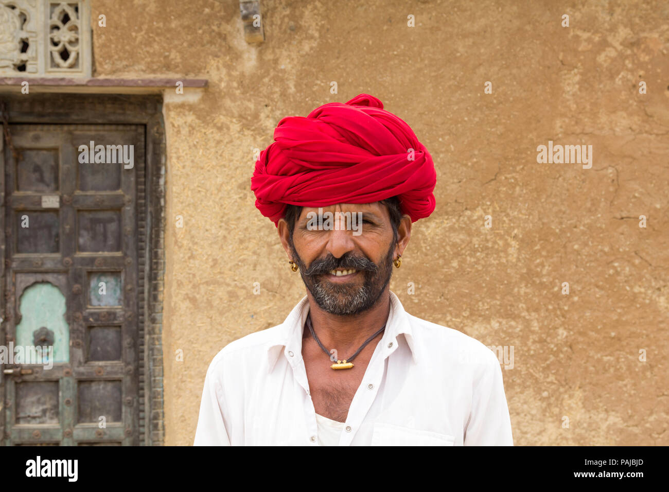 Uomo indiano con la barba che indossa un turbante rosso in piedi fuori da una casa rurale tradizionale a Shahpura, Rajasthan, India. Foto Stock