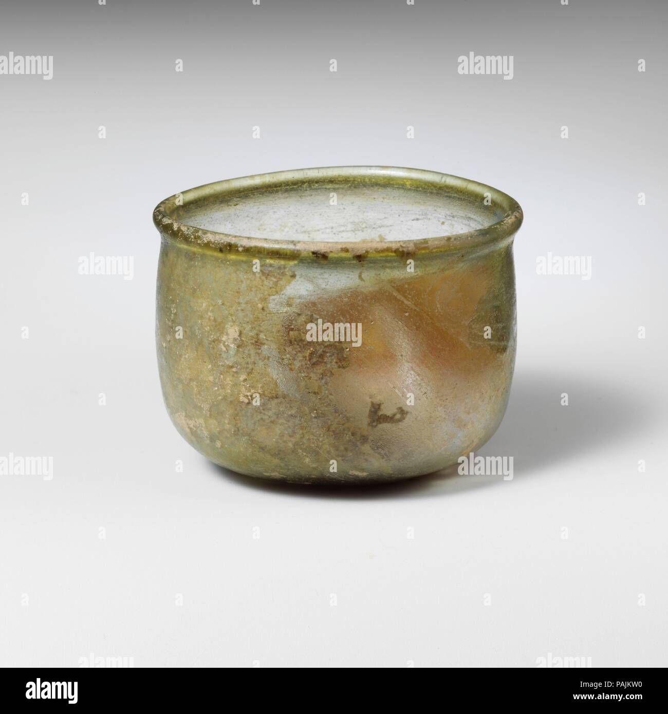 Bicchiere di vetro. Cultura: Romano. Dimensioni: 1 11/16 x 2 3/16a. (4,2 x 5,5 cm). Data: 2a-3a secolo D.C.. Traslucido giallo pallido verde. Arrotondati, addensato, e leggermente outsplayed rim; corpo cilindrico con lati leggermente convessi; fondo arrotondato con piccola e profonda kick al centro. Intatti; pinprick bolle e striature di soffiaggio; assopimento e iridescenza su esterno, cremosa sulla resistenza agli agenti atmosferici la maggior parte degli interni. Museo: Metropolitan Museum of Art di New York, Stati Uniti d'America. Foto Stock