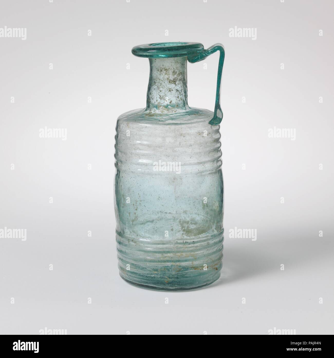 Il cilindro di vetro caraffa. Cultura: Romano. Dimensioni: 4 9/16a. (11.6cm). Data: 3° secolo D.C.. Blu traslucido verde; impugnatura in uno stesso colore. Cerchio tubolare, ripiegato e rotonda e in, e appiattito nella parte superiore; leggermente irregolare collo cilindrico; ampia, lievemente spalla spiovente; corpo cilindrico con lati leggermente convessi; fondo piatto, leggermente spinto in al centro, con piccolo giro pontil mark; largo e piatto maniglia a fascia attaccata al bordo superiore del corpo, redatto in senso verticale e successivamente trasformato in e verso il basso e trascinato sul lato inferiore del cerchio e la parte superiore del collo. Corpo soffiato in tre parti di stampo due sezioni verticali, e Foto Stock