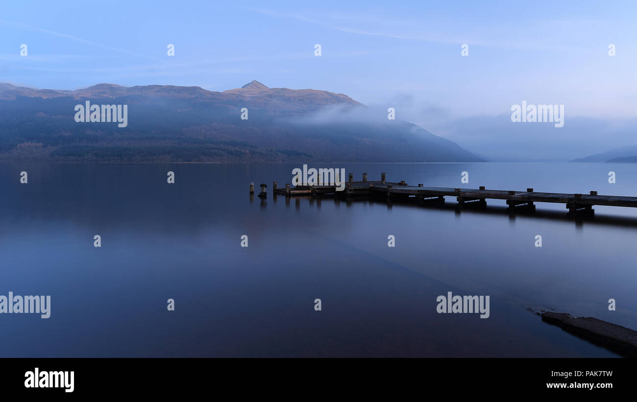 Misty Loch Lomond al crepuscolo quando l'ultima luce del sole ha colpito il pap di Ben Lomond sopra la foschia e Tarbet jetty Foto Stock