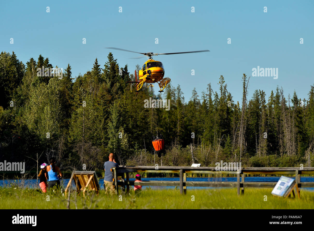 Un elicottero giallo carico di un secchio di acqua da un lago Alberta mentre la gente guarda Foto Stock