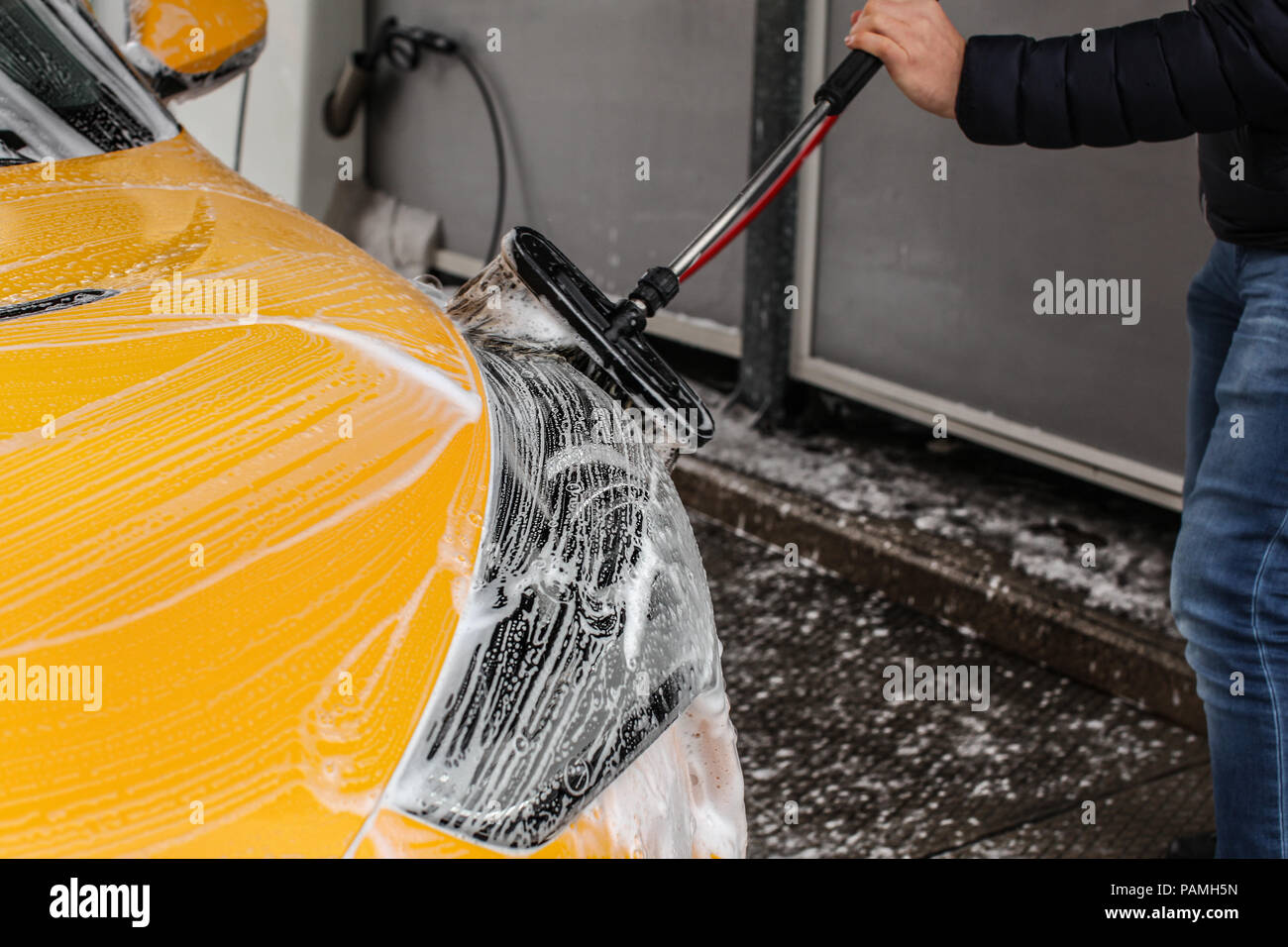 Uomo che pulisce la sua auto in self service carwash, spazzola lasciando tratti di shampoo sul faro anteriore. Foto Stock