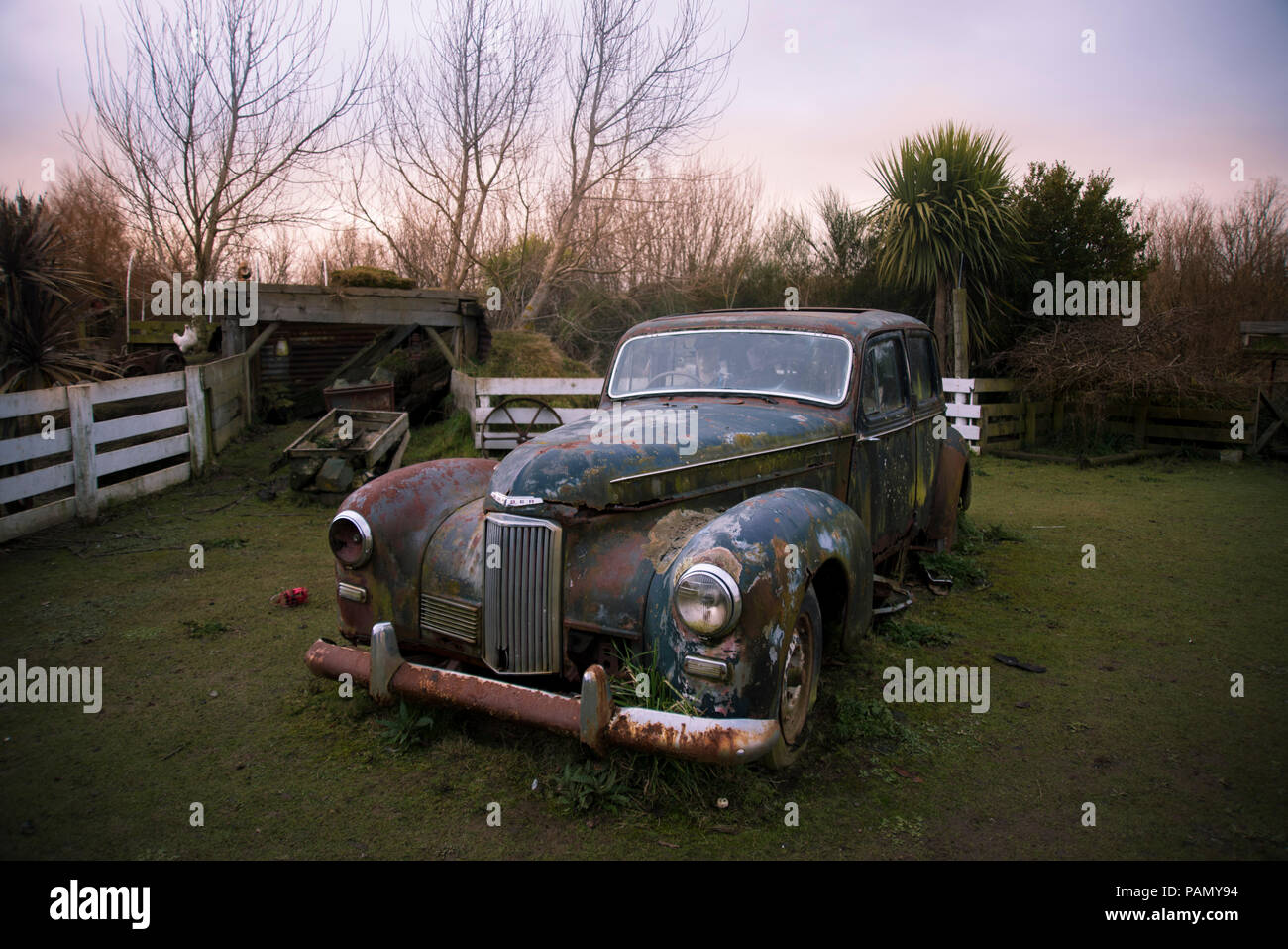 Vecchio arrugginito vintage Humber auto oltre un recinto di bianco, sull'erba in un cortile, durante il tramonto. Foto Stock