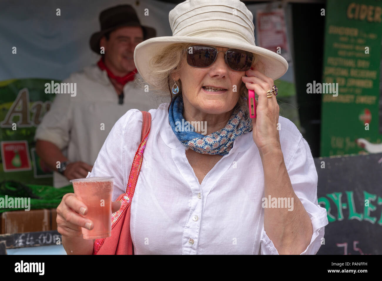 Donna matura indossando un cappello e occhiali da sole, tenendo in mano un drink e a parlare su un telefono mobile Foto Stock