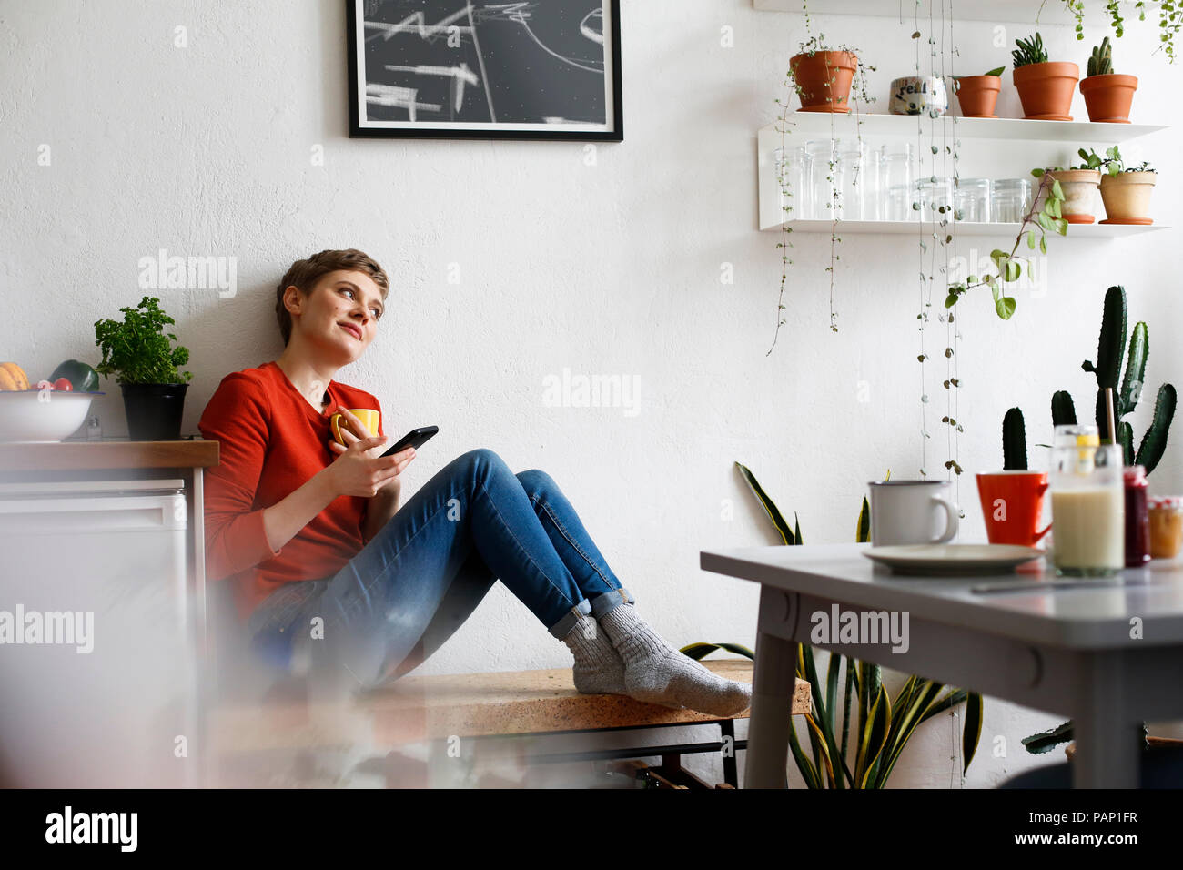 Donna seduta in cucina a bere caffè e controllare i messaggi sullo smartphone Foto Stock