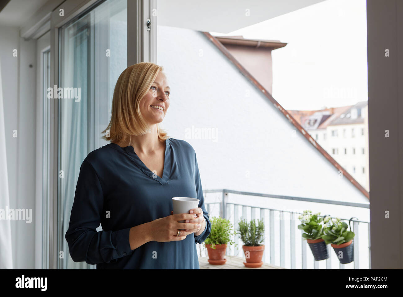 Donna sorridente holding tazza di caffè guardando fuori della porta del balcone Foto Stock