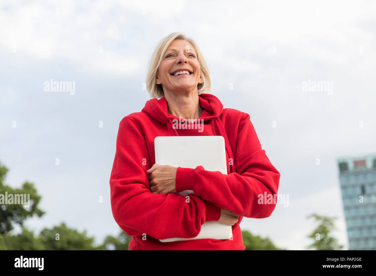 Felice senior donna che indossa felpa con cappuccio rosso azienda laptop all'aperto Foto Stock