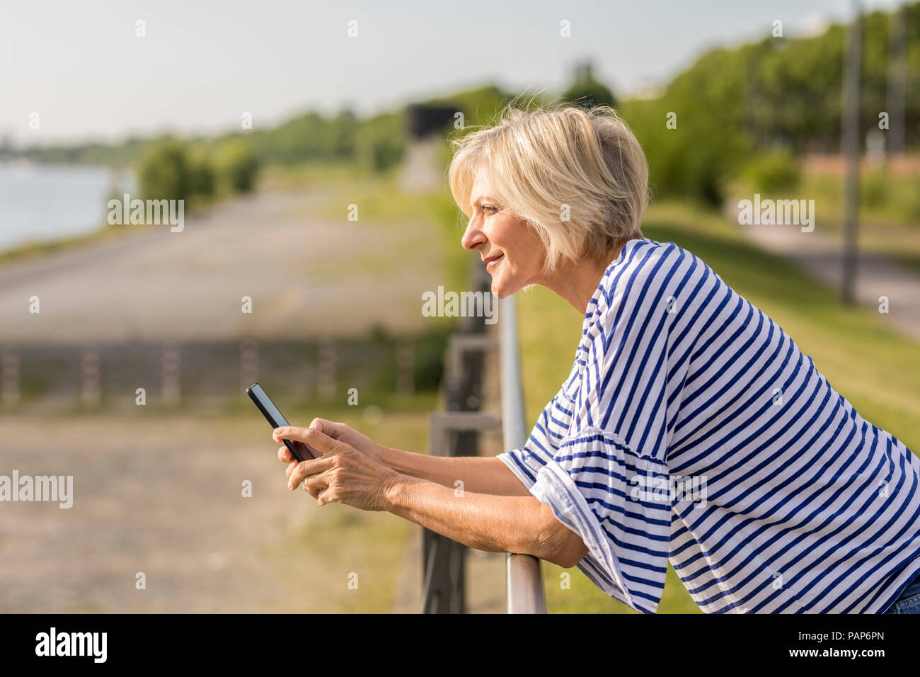 Sorridente donna senior con un telefono cellulare appoggiato sulla ringhiera a riverbank Foto Stock
