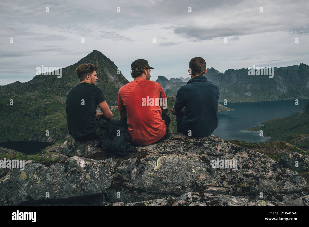 Norvegia Lofoten, Moskenesoy, tre giovani uomini seduti su una roccia, guardando sopra Kjerkefjord Foto Stock