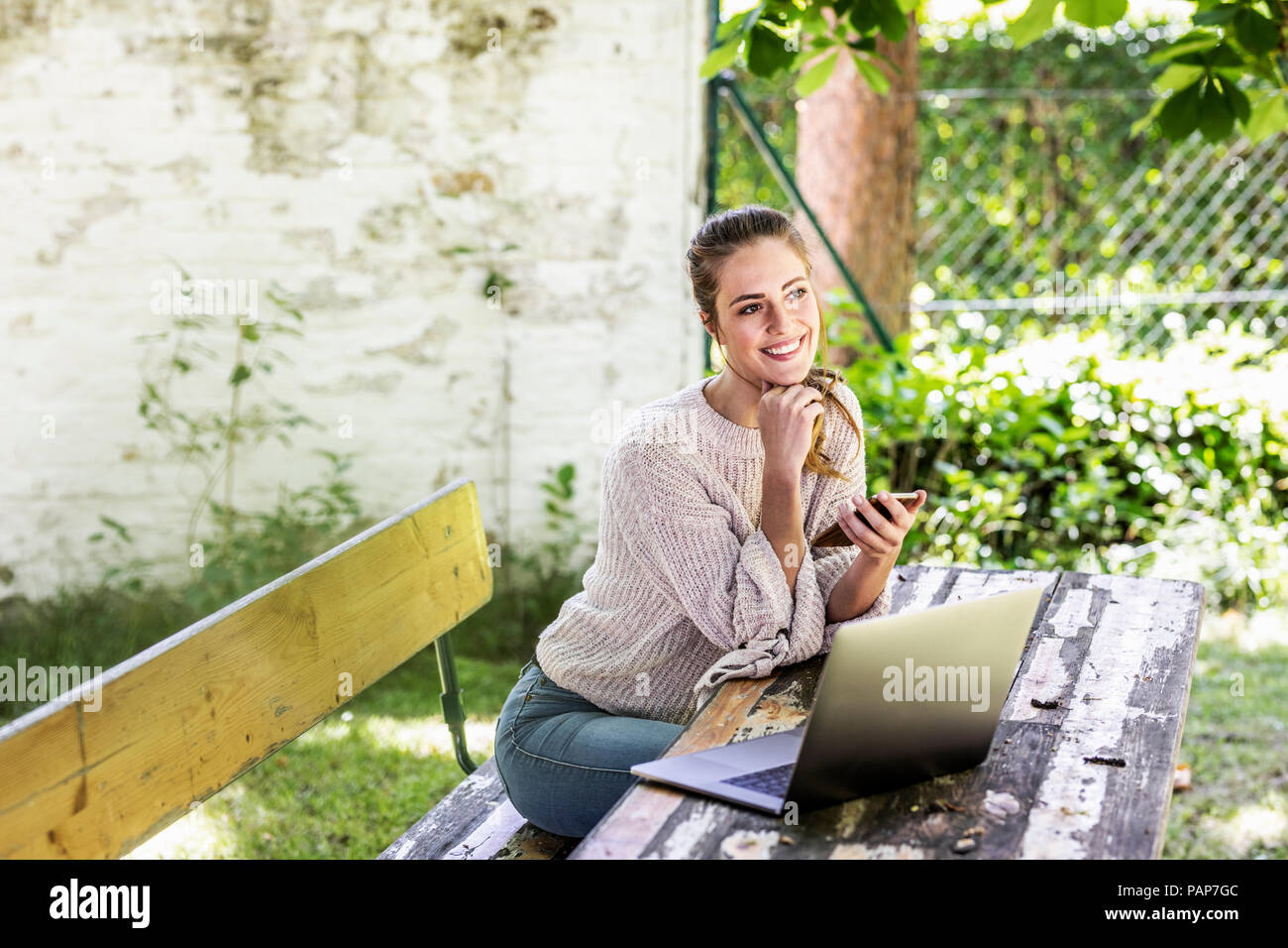 Felice fantasticando donna seduta in giardino con il laptop e telefono cellulare Foto Stock