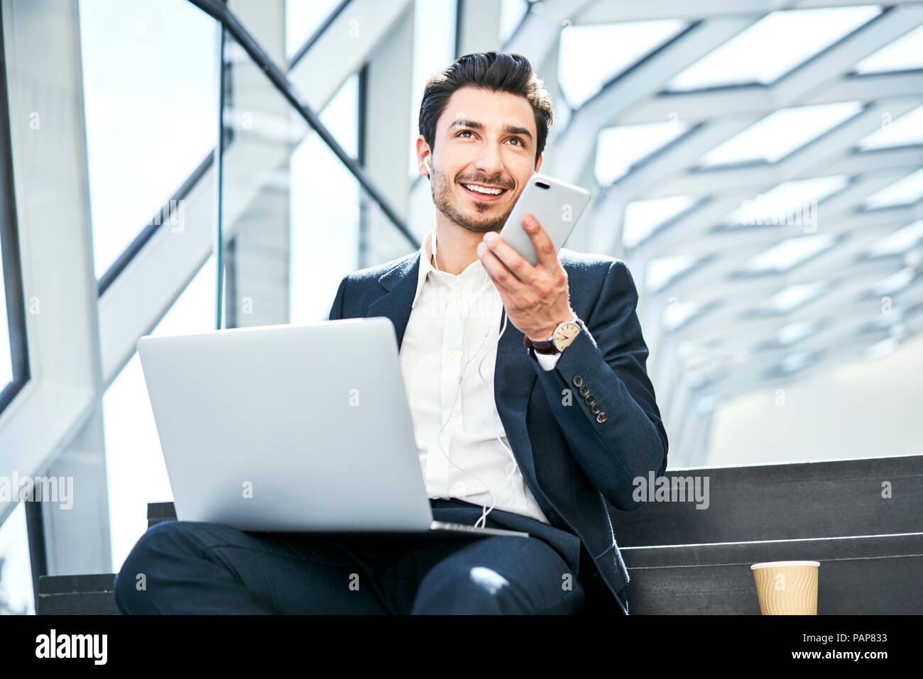 Imprenditore sorridente seduto su scale di indossare le cuffie con il telefono cellulare e il computer portatile Foto Stock