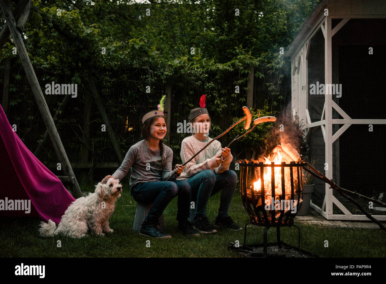 Due ragazze indossare copricapo di piume, grigliare salsicce oltre il campo di fuoco, dog sitter su prato Foto Stock
