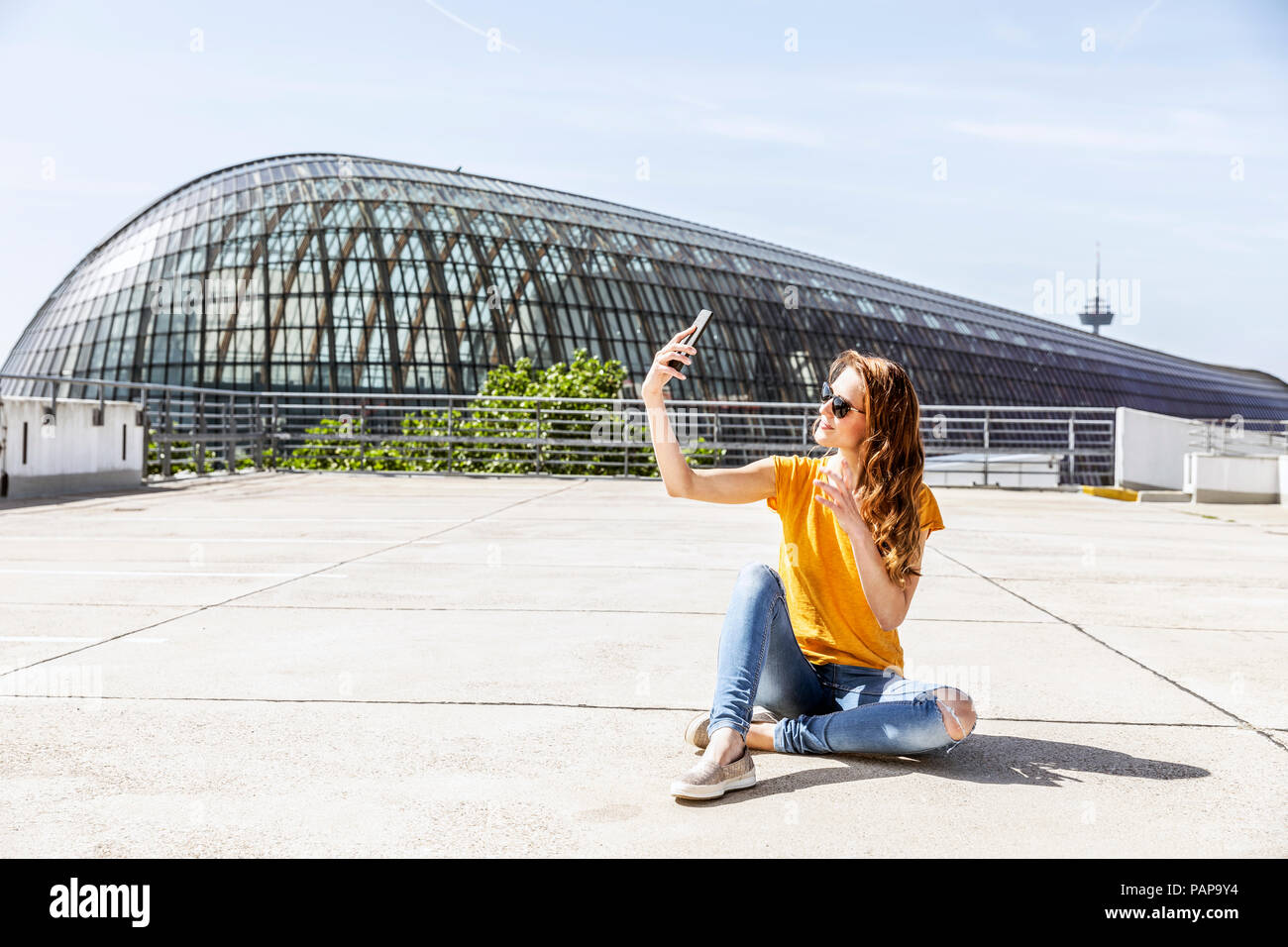 Germania, Colonia, sorridente donna seduta sul parcheggio livello tenendo selfie con lo smartphone Foto Stock