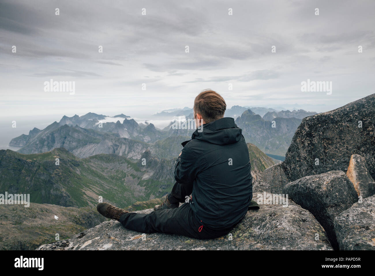 Norvegia Lofoten, Moskenesoy, giovane uomo seduto al Hermannsdalstinden, guardando sopra Kjerkefjord Foto Stock