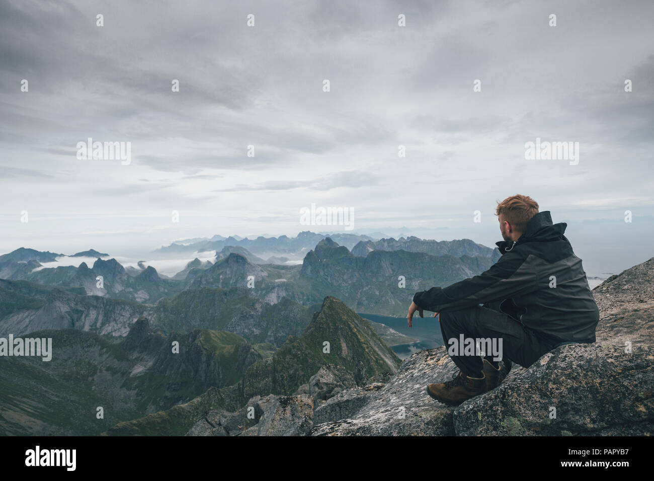 Norvegia Lofoten, Moskenesoy, giovane uomo seduto al Hermannsdalstinden, guardando sopra Kjerkefjord Foto Stock