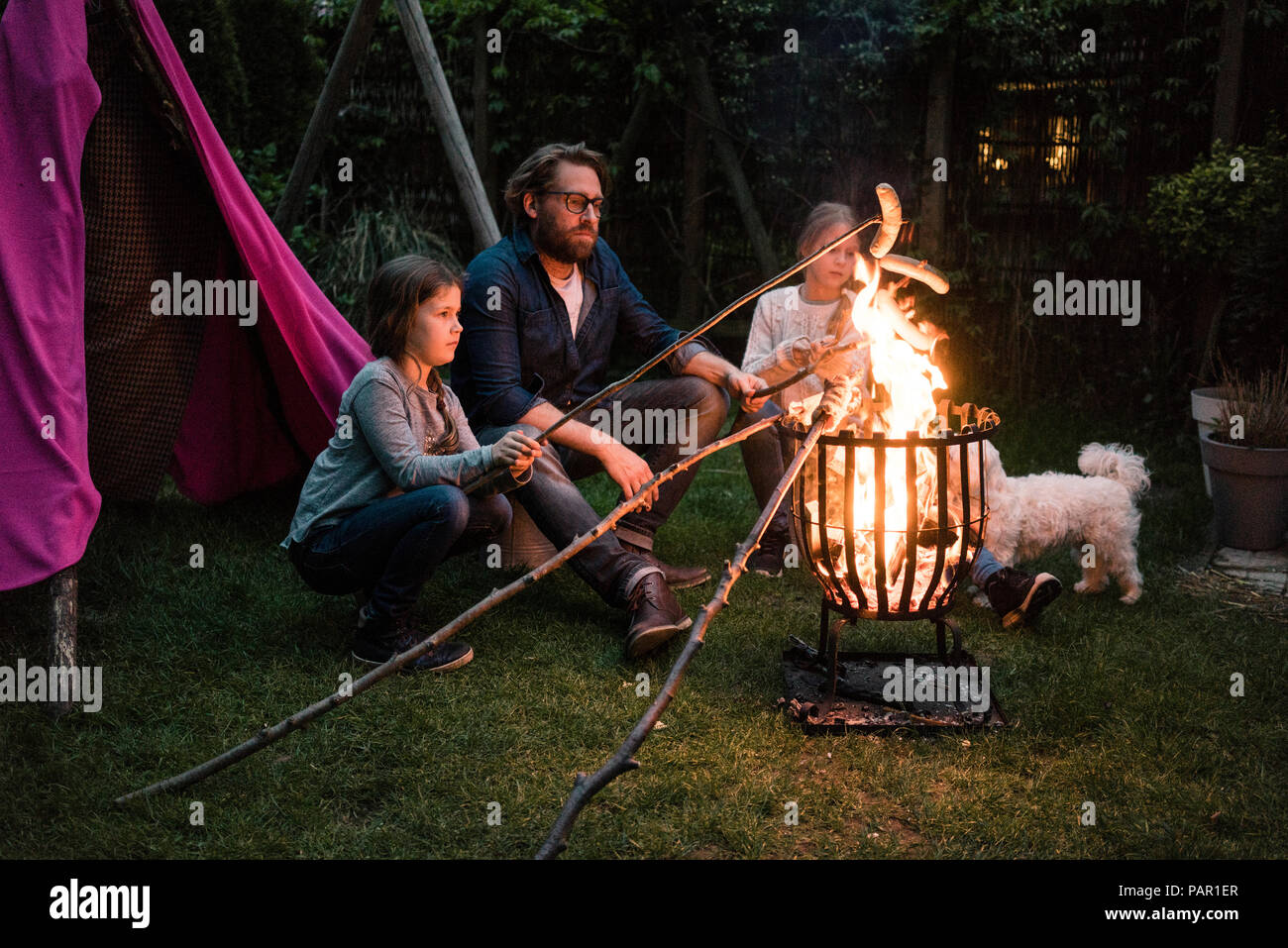 Uomo e due ragazze salsiccia cottura alla griglia su fuoco di campo in giardino Foto Stock