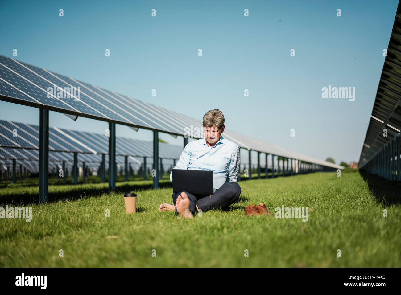 Coppia uomo seduto a piedi nudi sul prato, laptop e caffè di andare cup, impianto solare Foto Stock